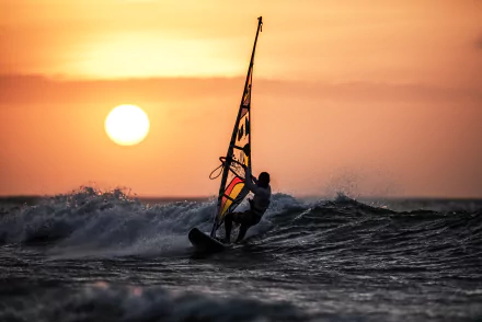 A windsurfer glides over ocean waves at sunset, with a vibrant sun casting a warm glow, showcasing the thrill of sports in this stunning 4K Ultra HD wallpaper.