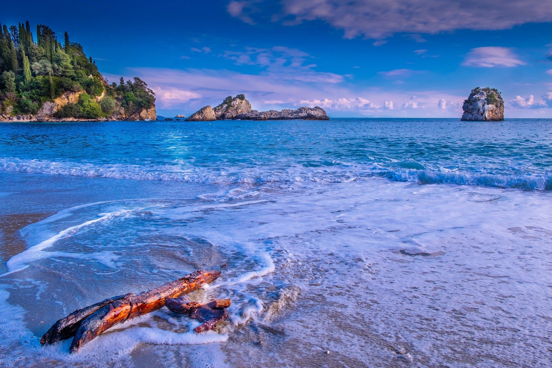 Scenic HD desktop wallpaper of a coastal ocean view featuring driftwood on a sandy shore, vibrant blue sea, and distant rocky outcrops under a partly cloudy sky.