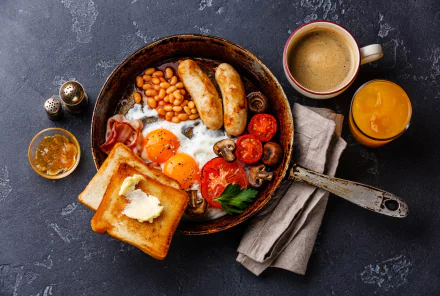 A beautifully arranged breakfast spread featuring toast, eggs, sausages, baked beans, tomatoes, and mushrooms, accompanied by coffee and juice, set against a dark background.
