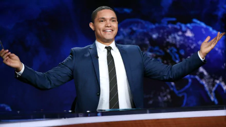 Trevor Noah smiles and gestures from the host's desk in this HD desktop wallpaper for The Daily Show with Trevor Noah, featuring a vibrant world map background.