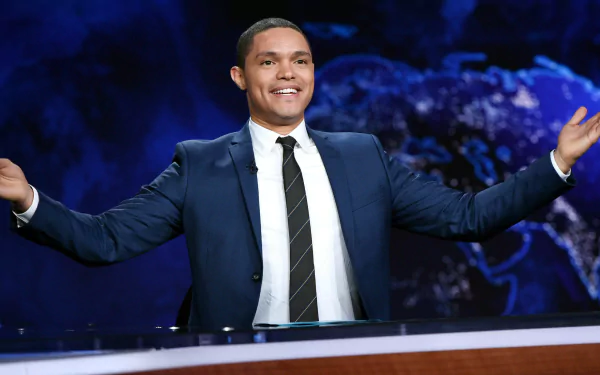 Trevor Noah smiles and gestures from the host's desk in this HD desktop wallpaper for The Daily Show with Trevor Noah, featuring a vibrant world map background.