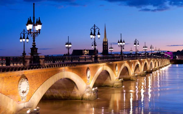4K Ultra HD wallpaper of a beautifully lit man-made bridge spanning calm water at dusk, featuring ornate street lamps and a church steeple in the background.