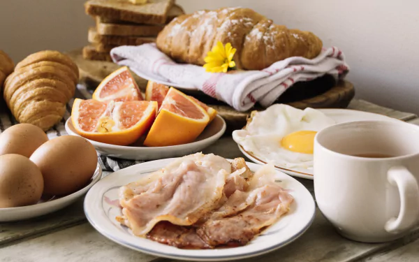 2K Quad HD PC desktop wallpaper/background showing a breakfast spread: croissant, bacon, fried egg, grapefruit wedges, whole eggs, sliced bread and a coffee cup on a tablecloth.