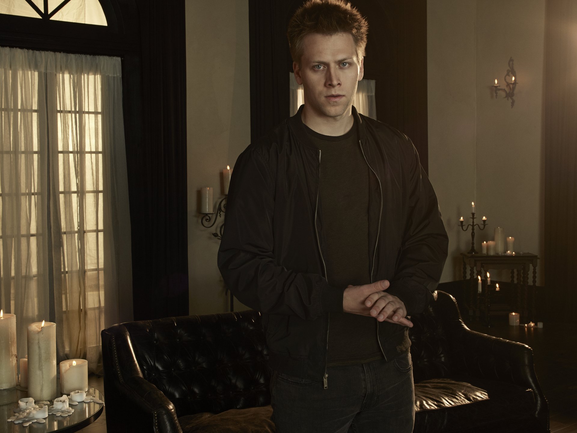 2K Quad HD PC desktop wallpaper: lone man in a dim, candlelit room with leather sofa and tall windows — Cloak & Dagger TV show background.