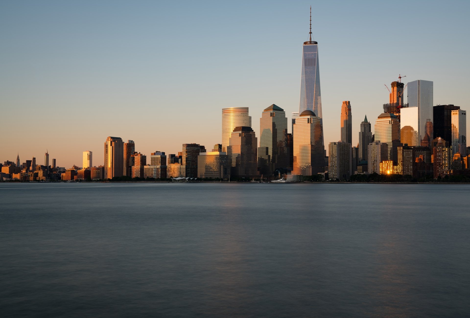 Sunset view of Manhattan skyscrapers, including the One World Trade Center, with the calm river in the foreground, captured in 8K Ultra HD resolution.