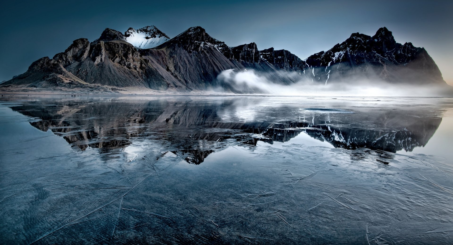 HD desktop wallpaper showing icy mountain peaks reflected on a still, frozen lake under a moody sky, capturing the serene beauty of nature.