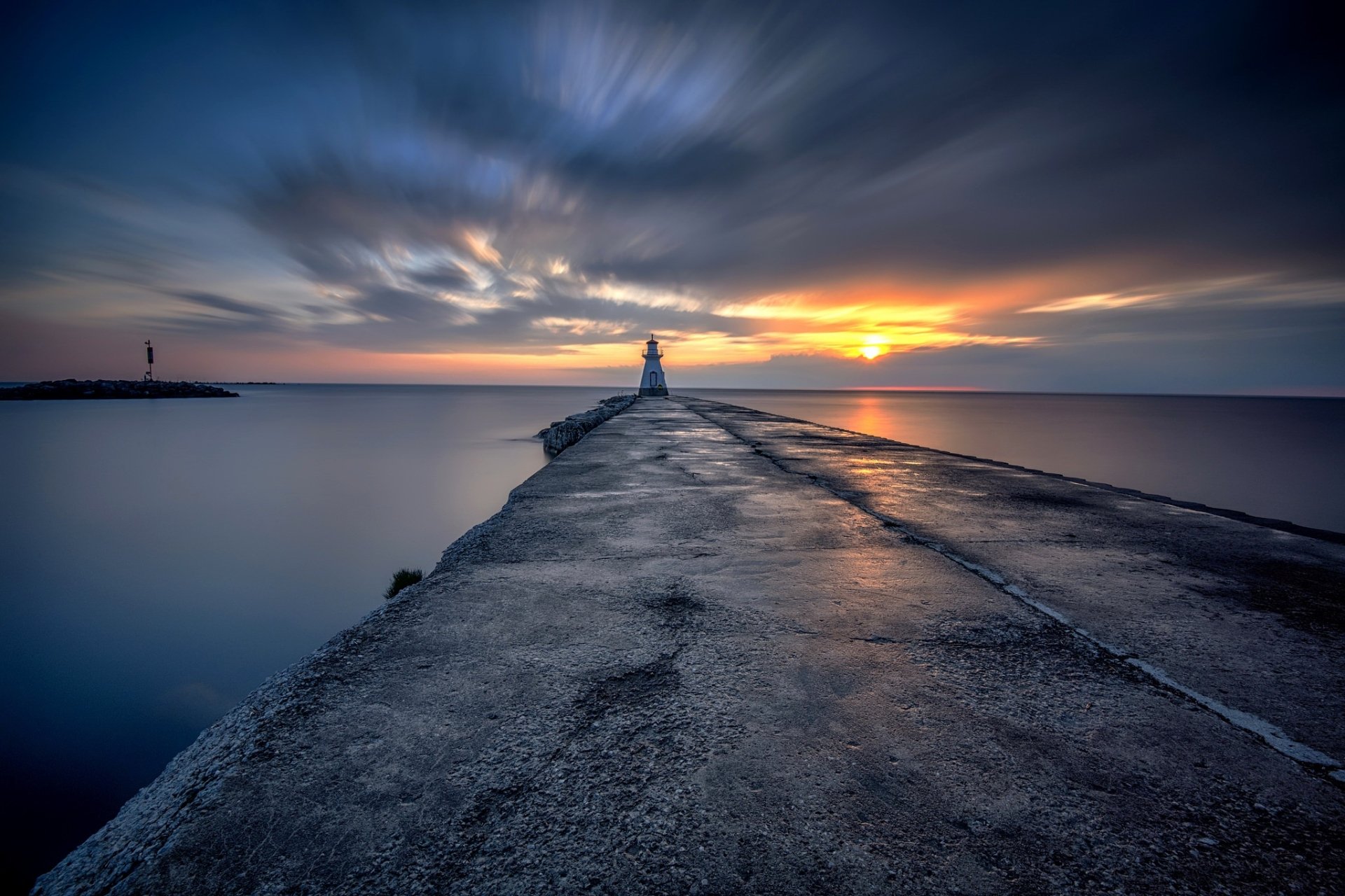 HD desktop wallpaper of a man-made pier leading to a lighthouse at sunset, with the ocean stretching to the horizon under a dramatic sky.