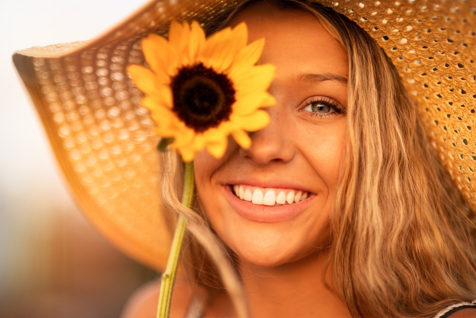 HD PC desktop wallpaper: smiling blonde woman model in a straw hat, sunflower covering one eye, bright close-up of her face.
