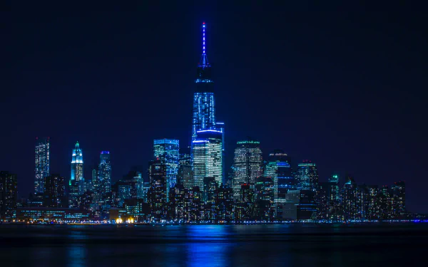 Night view of Manhattan skyscrapers in New York, illuminated in blue lights, reflecting on the water in this 8K Ultra HD cityscape wallpaper.