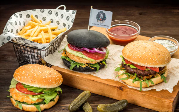 HD desktop wallpaper featuring a still life of three burgers, a basket of French fries, pickles, and dipping sauces arranged on a wooden surface.