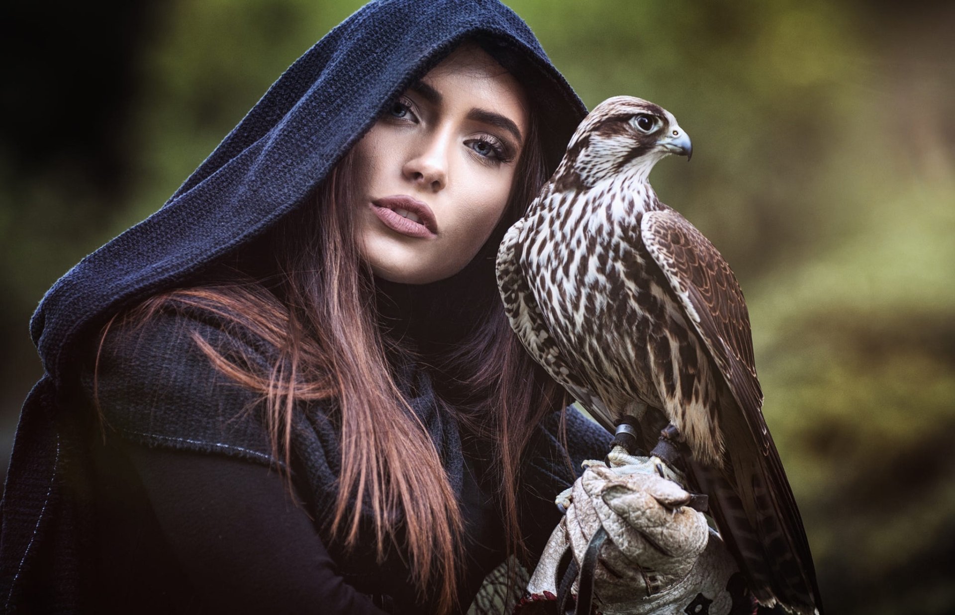 A brunette model with striking blue eyes wearing a dark hood holds a majestic falcon, set against a blurred natural background in this HD desktop wallpaper.