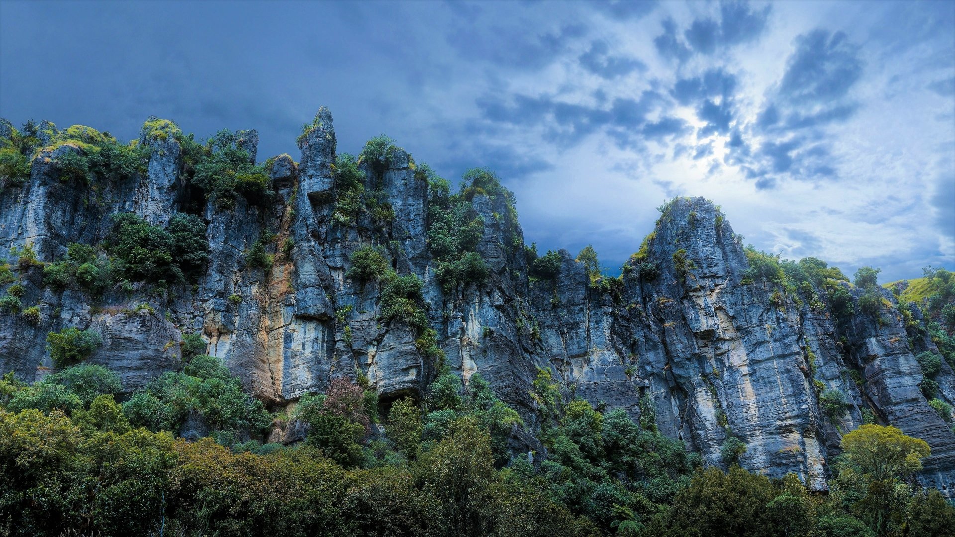 4K Ultra HD image of a rugged New Zealand mountain cliff with lush greenery under a cloudy sky, showcasing dramatic natural landscape.