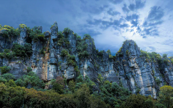 4K Ultra HD image of a rugged New Zealand mountain cliff with lush greenery under a cloudy sky, showcasing dramatic natural landscape.
