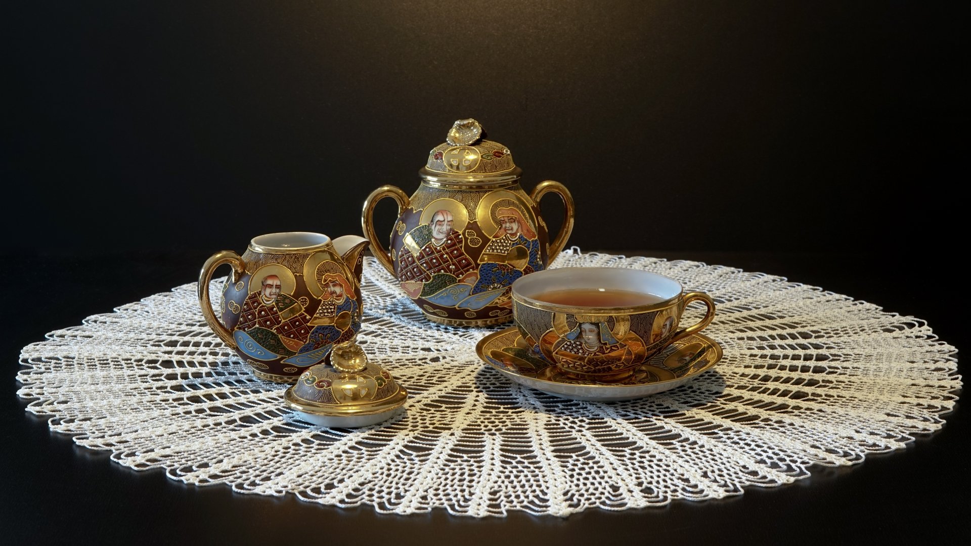A 4K Ultra HD still life of an ornate teapot, creamer, and teacup filled with tea, all placed on a lace doily against a dark background.