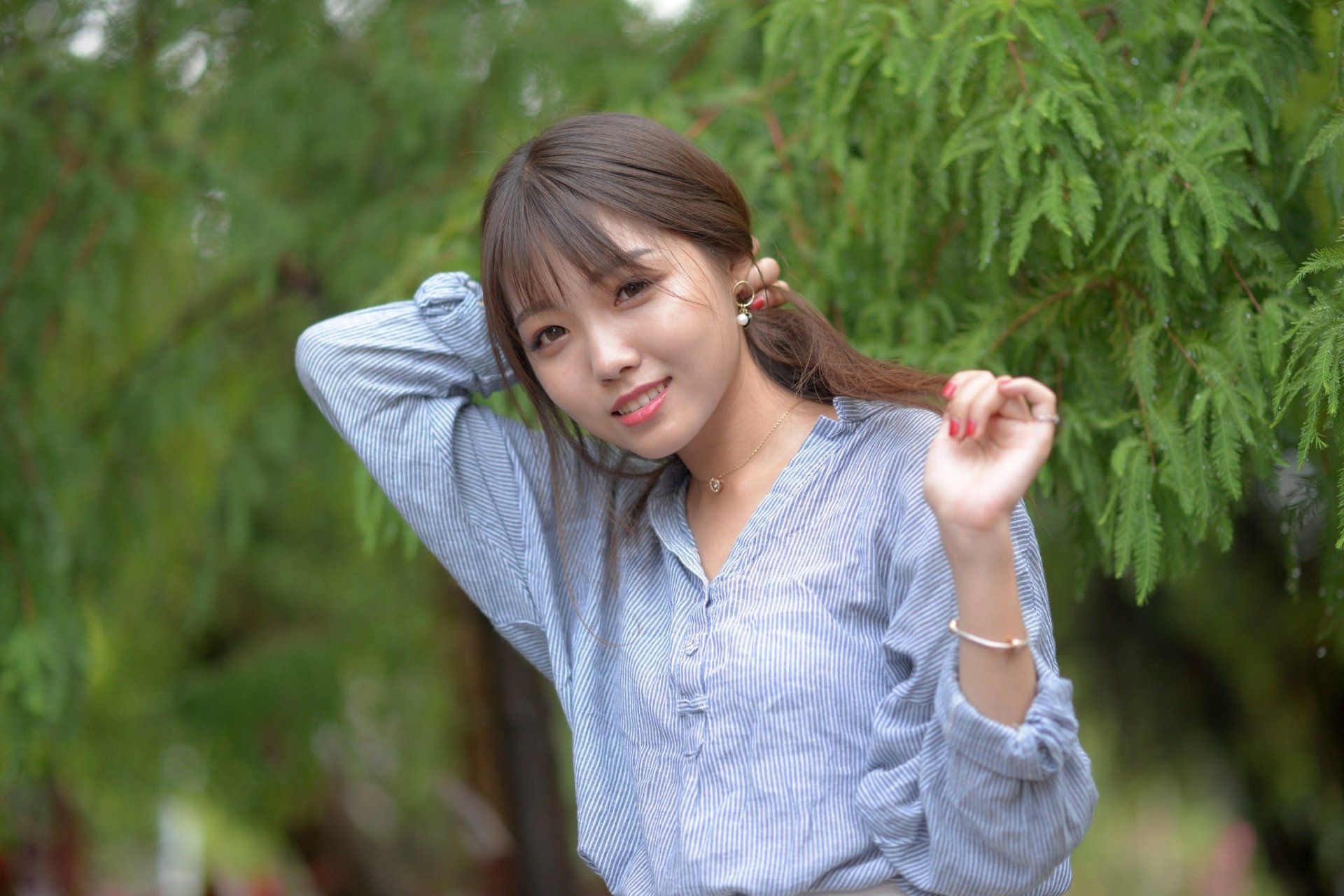 A smiling brunette woman with brown eyes, wearing a blue shirt, poses with her hand in her hair against a green, blurred background. The depth of field highlights her features. HD desktop wallpaper.