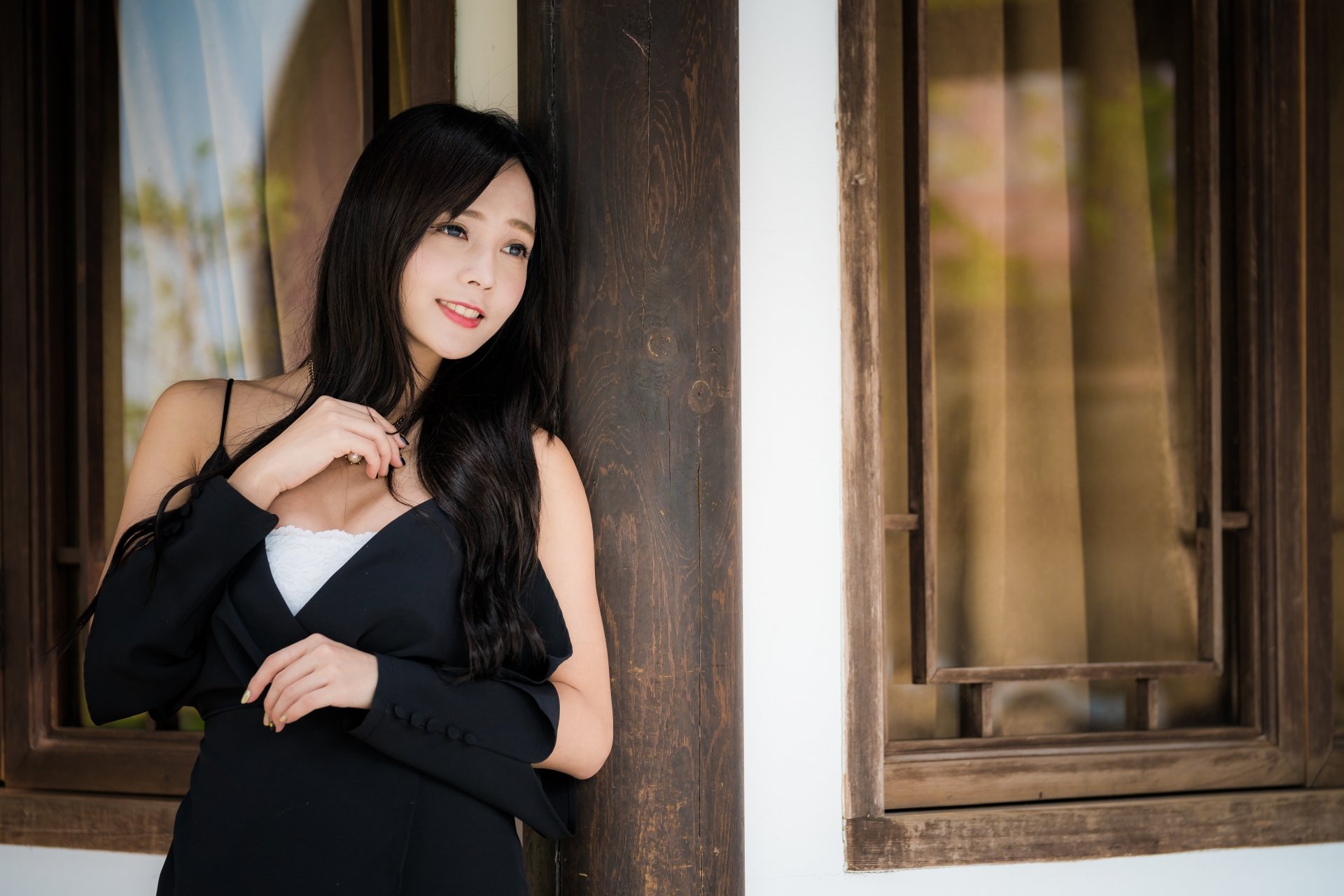 A smiling Asian woman with black hair stands by a wooden post, wearing a dark outfit. The HD desktop wallpaper captures her in a warm, natural setting.