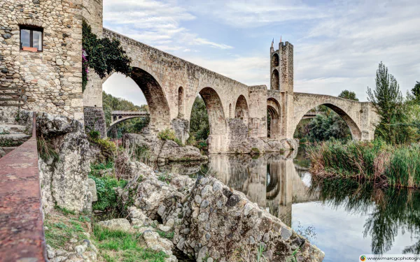 Scenic 4K Ultra HD view of the historic man-made stone bridge in Besalú, Catalonia, reflecting over the calm river with surrounding greenery and medieval architecture.