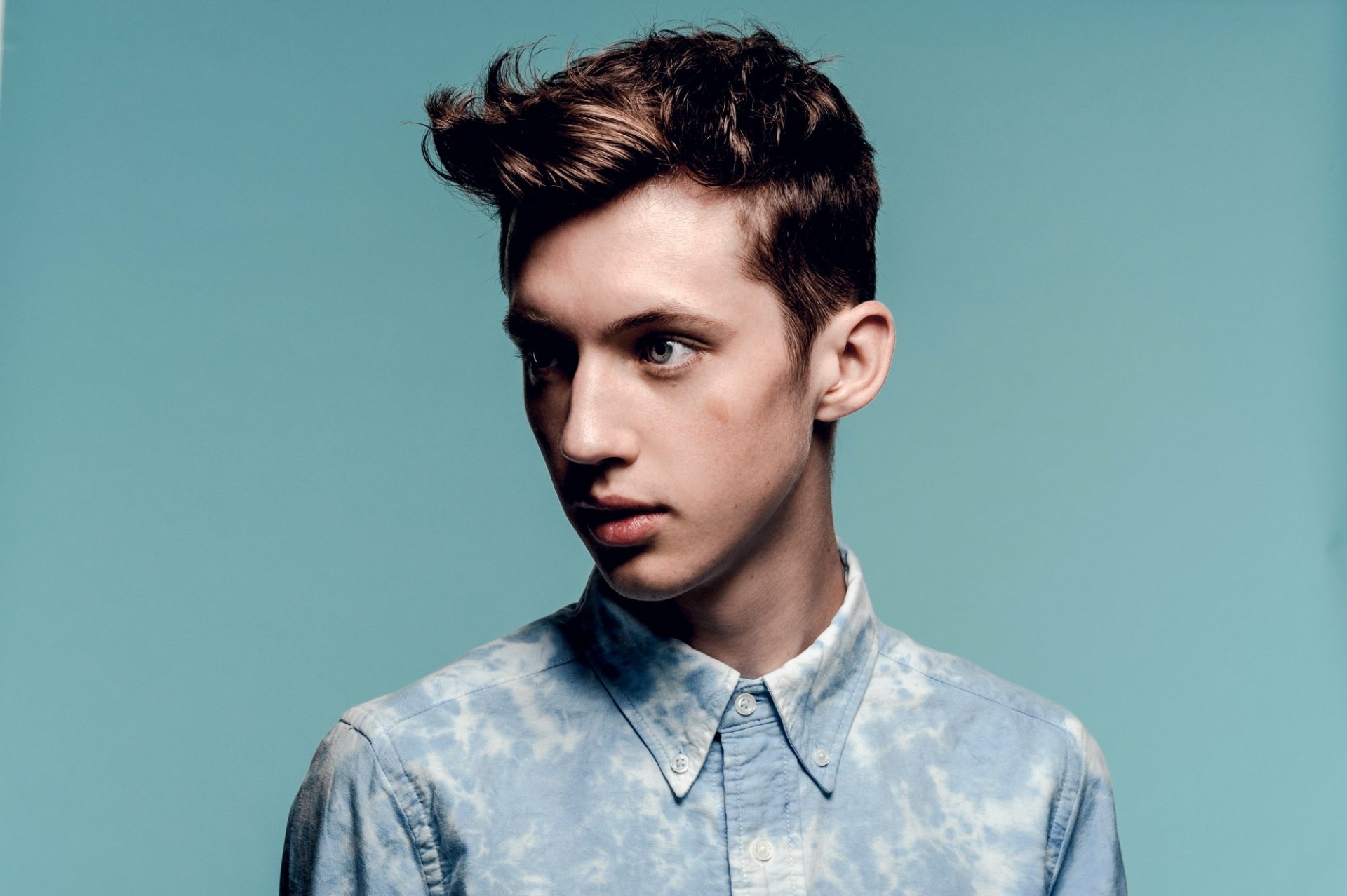HD desktop wallpaper featuring a portrait of a young man with stylish hair and a blue patterned shirt against a teal background.