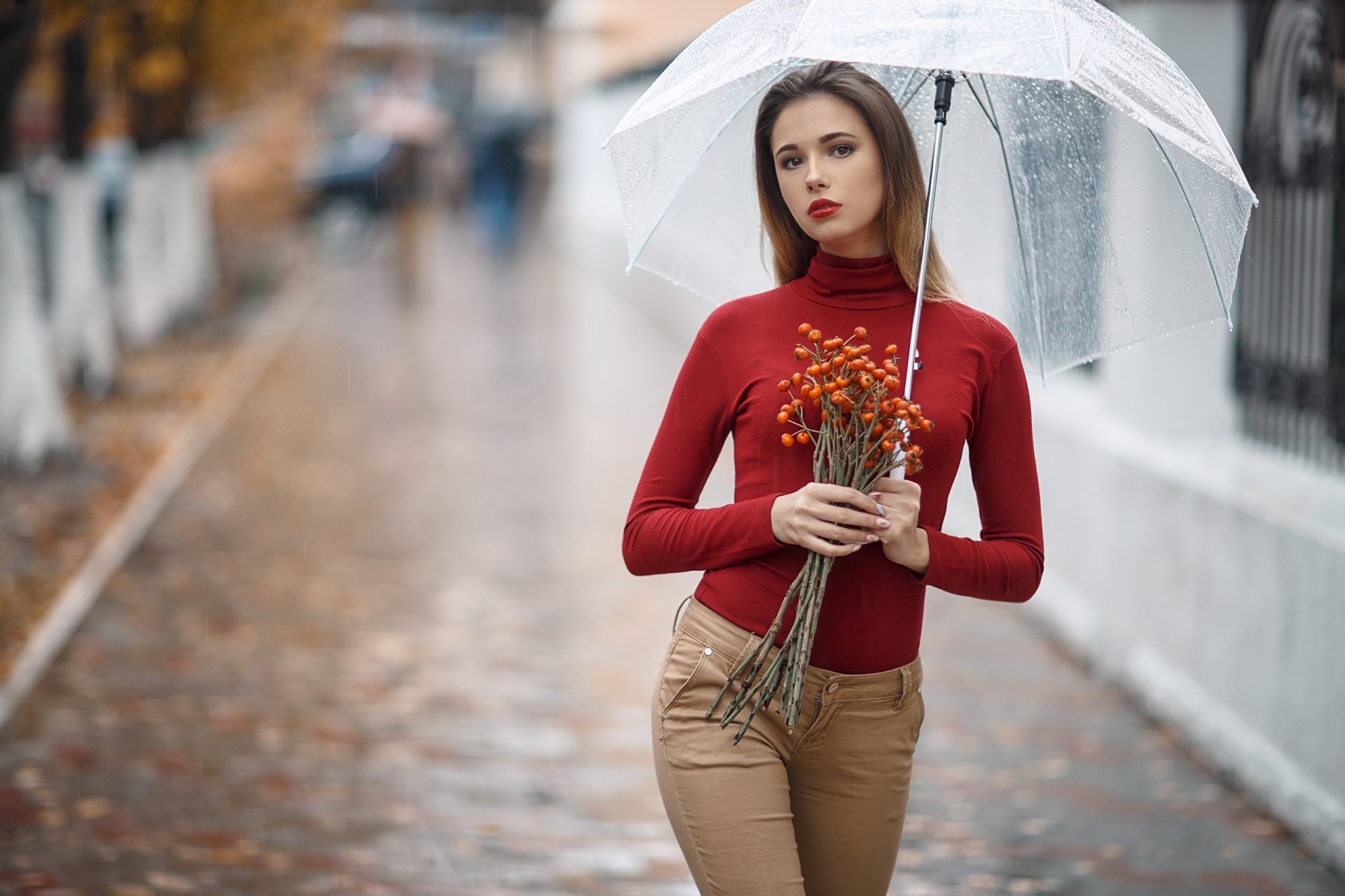 Brunette model with brown eyes and red lipstick holds a clear umbrella and small bouquet on a rainy street; shallow depth-of-field, 2K Quad HD PC desktop wallpaper/background.