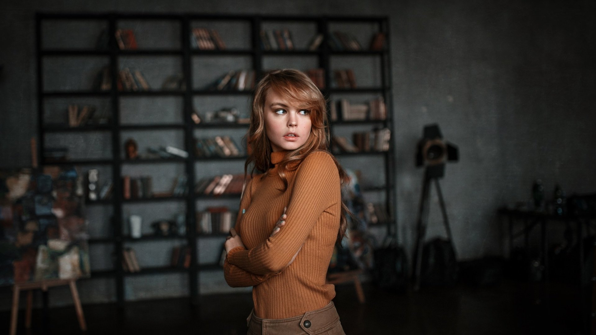 HD desktop wallpaper featuring Russian model Anastasiya Scheglova in a brown turtleneck, standing with arms crossed in an industrial-style room with shelves and camera equipment.