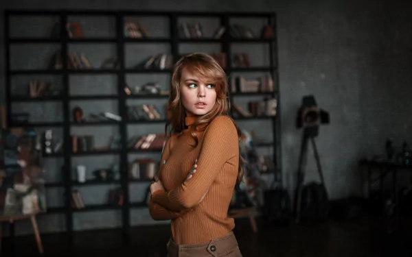 HD desktop wallpaper featuring Russian model Anastasiya Scheglova in a brown turtleneck, standing with arms crossed in an industrial-style room with shelves and camera equipment.
