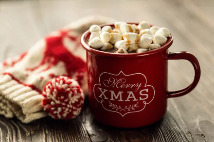 A red mug filled with hot chocolate topped with marshmallows, featuring a Merry Xmas message, set beside knitted winter accessories on a wooden surface.