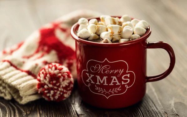 A red mug filled with hot chocolate topped with marshmallows, featuring a Merry Xmas message, set beside knitted winter accessories on a wooden surface.