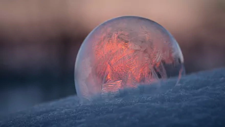 HD desktop wallpaper of a frozen ice bubble resting on snow, glowing softly with warm light against a blurred natural background.