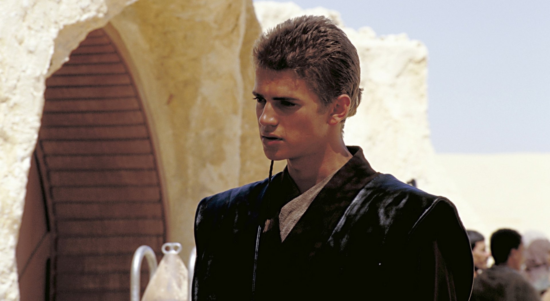 HD movie desktop wallpaper: young man in a dark robe standing in a sunlit desert courtyard, cinematic still evoking a sci‑fi prequel.
