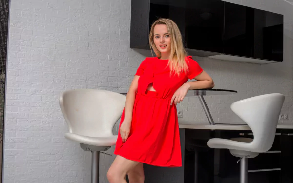 Blonde woman model in a vibrant red dress smiles confidently, posing indoors by modern white bar stools against a white brick wall—a sharp HD desktop wallpaper scene.