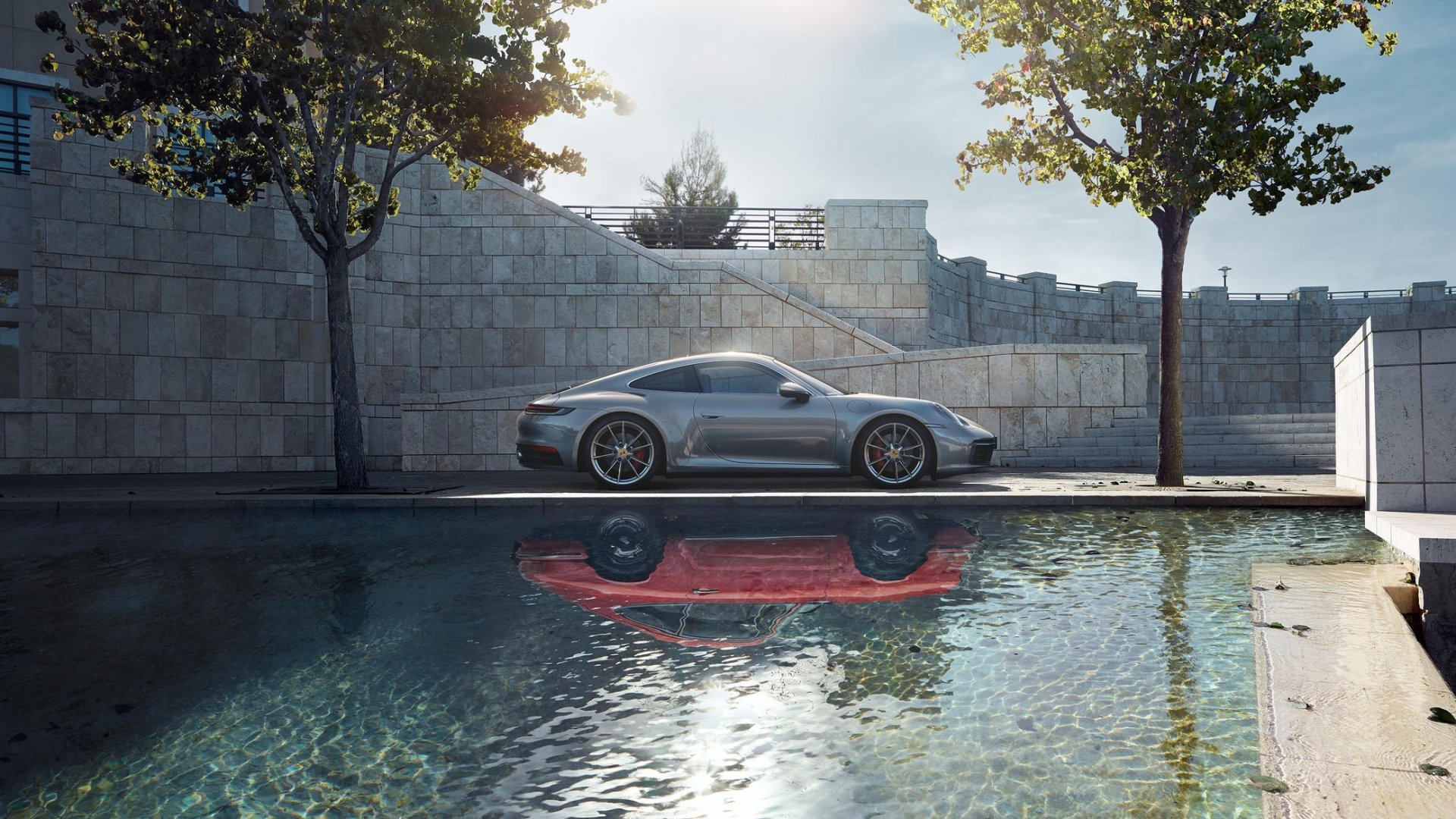 Silver Porsche 911 Carrera parked beside a reflecting pool with modern stone walls and trees — HD PC desktop wallpaper background.