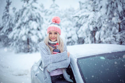 Smiling blonde woman with blue eyes in a pink pom-pom hat and gray sweater by a snow-covered car amid falling snow — HD winter desktop wallpaper.