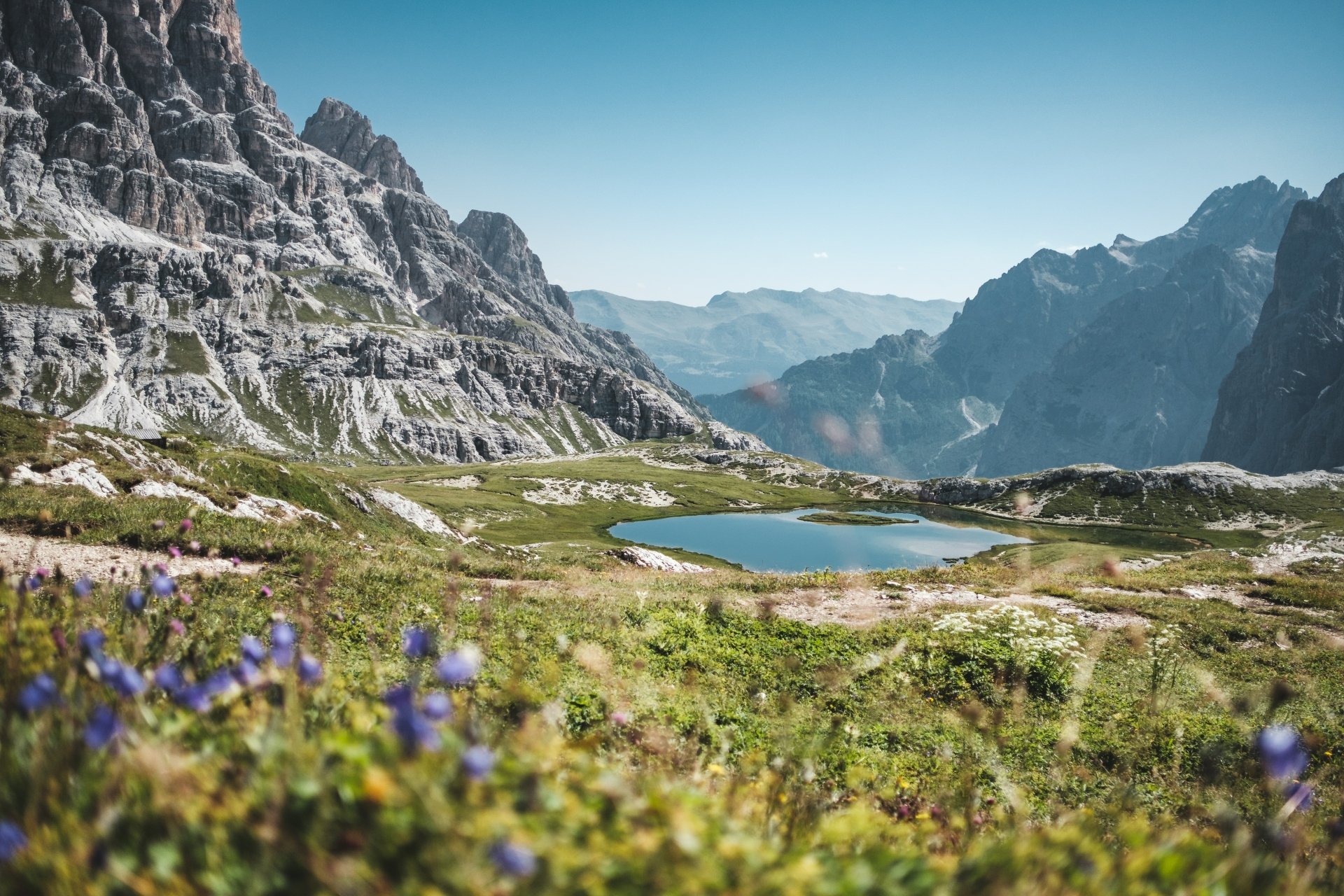 4K Ultra HD desktop wallpaper showing a serene mountain landscape with clear blue sky, rocky peaks, a peaceful pond, and vibrant wildflowers in the foreground.