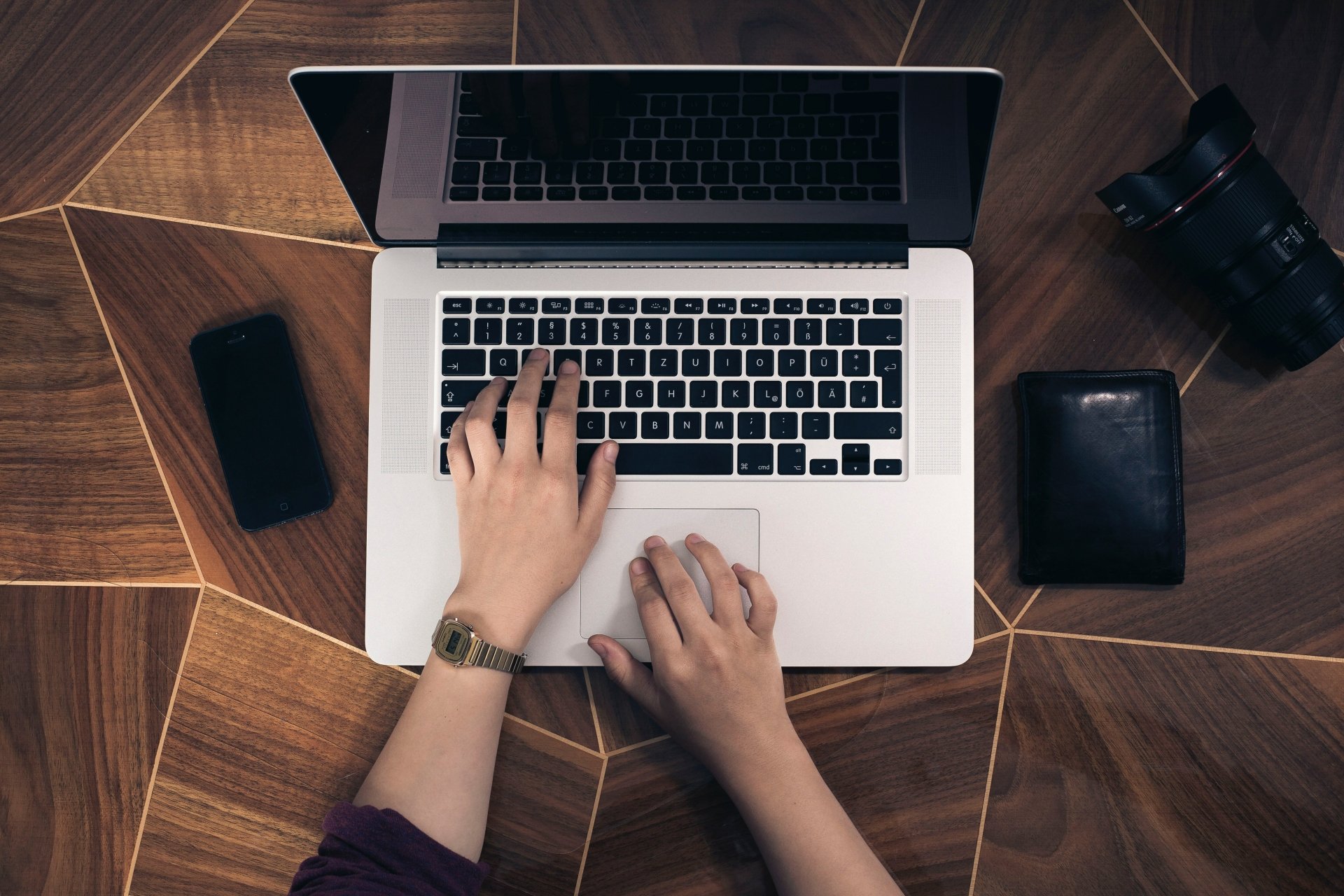 4K Ultra HD PC desktop wallpaper showing top-down view of a laptop with hands typing, a smartphone, camera, and wallet on a geometric wooden surface — technology, laptop