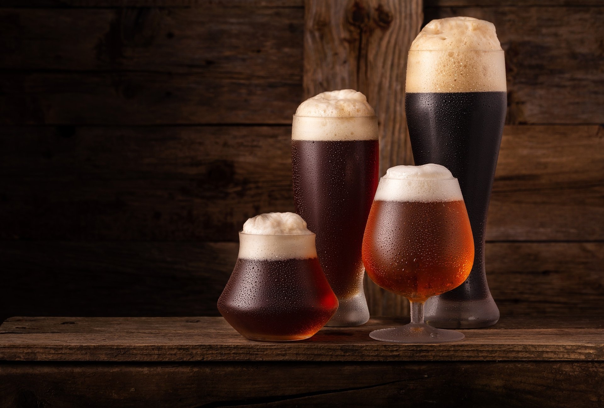 HD PC desktop wallpaper showing four glasses of beer with frothy heads on a wooden surface against a dark rustic background, representing alcohol, drink, and food.