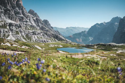 4K Ultra HD desktop wallpaper showing a serene mountain landscape with clear blue sky, rocky peaks, a peaceful pond, and vibrant wildflowers in the foreground.
