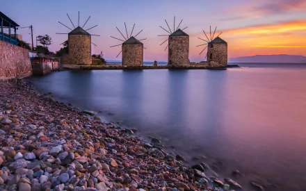 Four traditional Greek windmills line the rocky shore of the Aegean Sea at sunset, captured in a vivid HD desktop wallpaper scene.