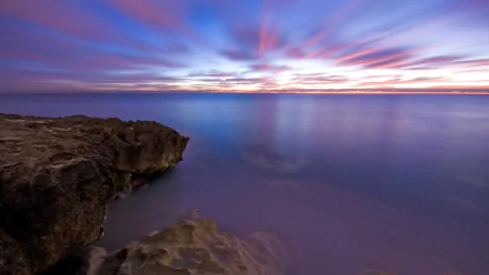 HD desktop wallpaper showcasing a serene ocean scene at morning with rocky cliffs under a purple and blue sky.