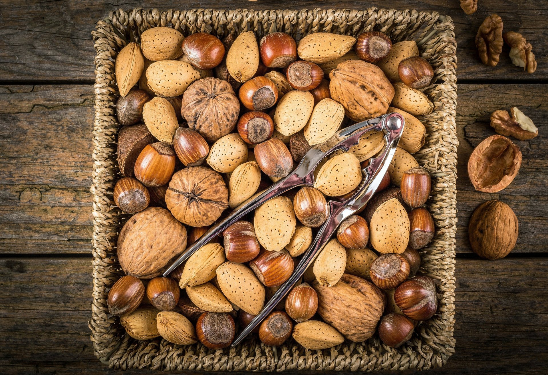 HD desktop wallpaper showing a variety of whole nuts in a basket with a metal nutcracker on top, set against a rustic wooden background.