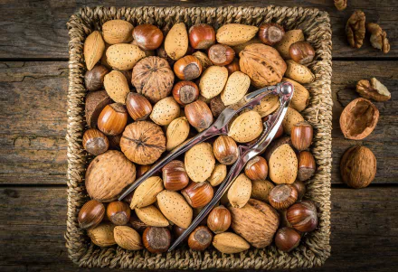 HD desktop wallpaper showing a variety of whole nuts in a basket with a metal nutcracker on top, set against a rustic wooden background.