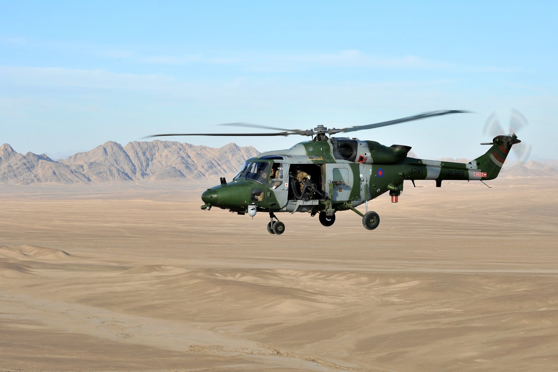 HD desktop wallpaper of a military Westland Lynx helicopter flying over a desert landscape with mountains in the background.