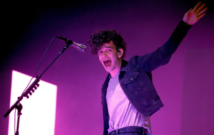 HD desktop wallpaper featuring a member of The 1975 performing onstage with a microphone, highlighted by purple lighting.